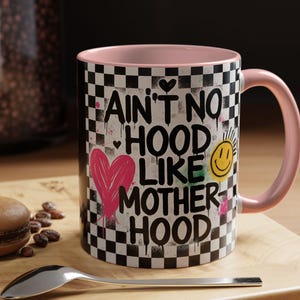 May include: A ceramic coffee mug with a pink interior and handle. The mug features a black and white chequered pattern with the words "AIN'T NO HOOD LIKE MOTHER HOOD" in a graffiti-style design, along with a pink heart and a smiling sun.