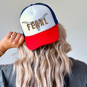 Op de afbeelding: Een rood, wit en blauwe trucker-pet met een wit voorpaneel met een bruine en zwarte luipaardprint stieren schedel met het woord "FEWAL" gedrukt in bruine letters.