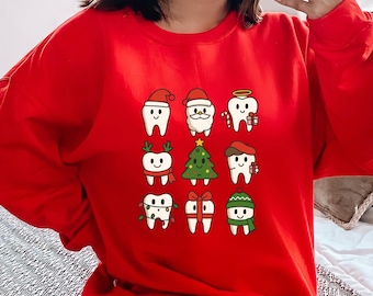 以下が含まれることがあります： クリスマスをテーマにしたデザインが施された、9つの漫画の歯が格子状に配置された赤いスウェットシャツ。歯にはサンタ帽、ハロー、スカーフが描かれており、キャンディケイン、クリスマスツリー、プレゼントで飾られています。