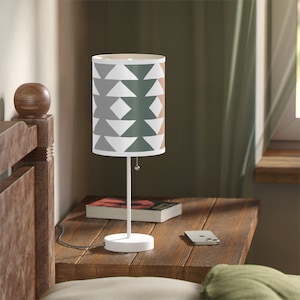May include: A white table lamp with a geometric patterned lampshade in shades of gray, green, and peach. The lamp is on a wooden table with a book and a phone.