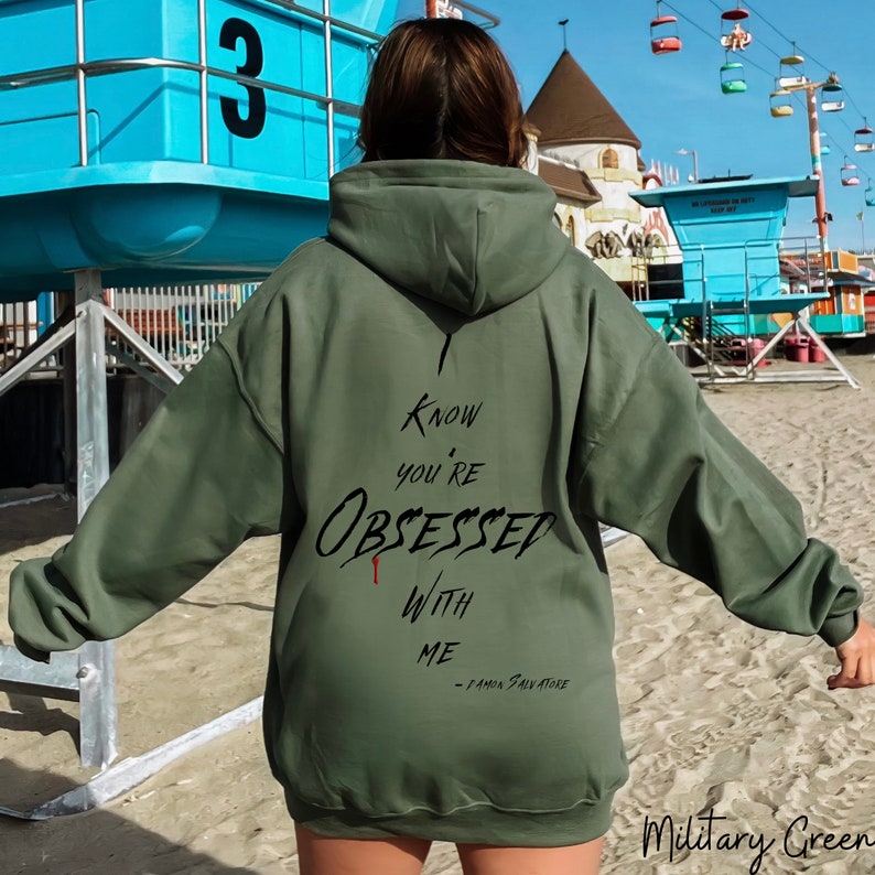 Pode incluir: Uma camisola de capuz verde com o texto "I know you're obsessed with me - Damon Salvatore" impresso nas costas. A camisola &eacute; de cor verde militar.