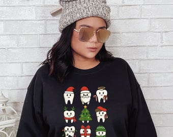 以下が含まれることがあります： クリスマスをテーマにした歯のデザインが施された黒いスウェットシャツ。サンタの帽子やスカーフ、クリスマスツリーなど、お祝いのアクセサリーを身につけた漫画の歯が描かれています。スウェットシャツは、リラックスしたカジュアルなスタイルです。