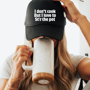 Puede incluir: Gorra de camionero negra con el texto blanco "I don't cook But I love to Stir the pot". Una persona sostiene una taza blanca con base de madera. La taza tiene motas negras.