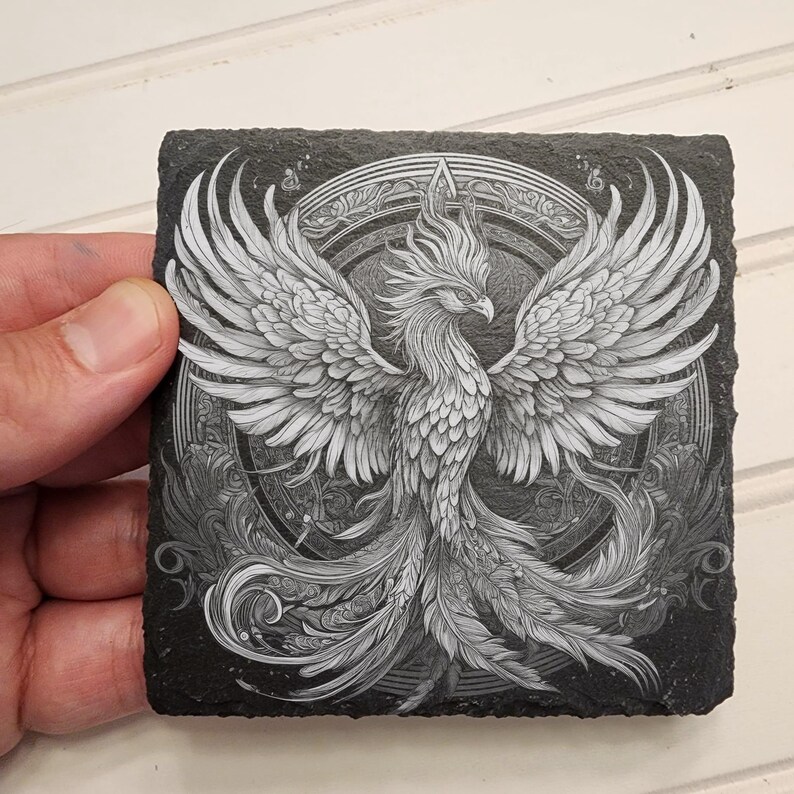 May include: Black slate coaster with a white detailed illustration of a phoenix with its wings spread. The phoenix is surrounded by a circular design.
