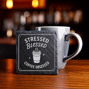 May include: A square, dark gray slate coaster with white text that reads "Stressed Blessed Coffee Obsessed" around a coffee cup graphic. A gray mug sits behind the coaster on a wooden surface.