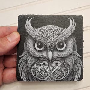 K&ouml;nnte beinhalten: Ein schwarzer Schieferuntersetzer mit einem detaillierten, wei&szlig;en Grafikdesign eines Eulen mit komplizierten keltischen Knotenmustern. Die Augen der Eule sind gro&szlig; und rund, und ihre Federn sind mit feinen Linien dargestellt.