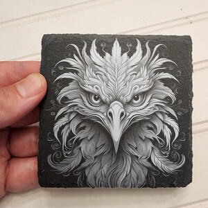 May include: A black slate coaster with a white and gray detailed illustration of an eagle's head. The eagle's eyes are wide open and its beak is pointed.