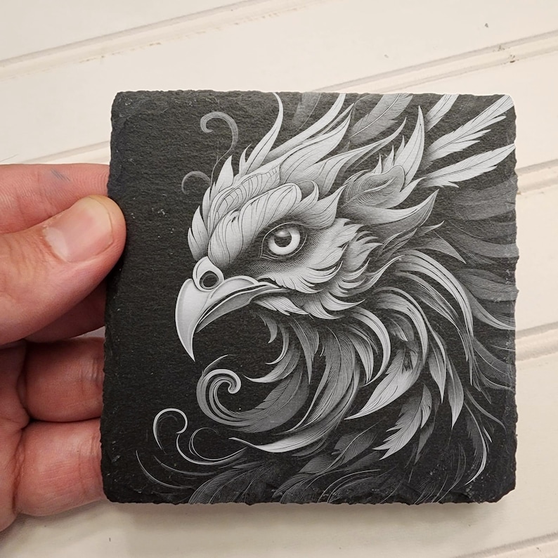 May include: Black and white detailed illustration of a bird of prey with intricate feathers and a sharp beak, printed on a black slate coaster.