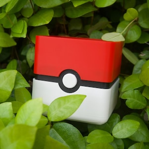 May include: A red, white, and black rectangular container, resembling a Poké Ball, sits amidst green foliage. The top half is red, the bottom white, with a black band and a white circle in the center.