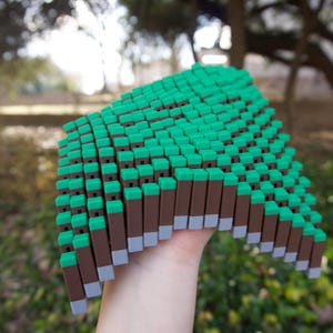 May include: A hand holds a pixelated object in green and brown, reminiscent of a tree or building. The object is made of many small, rectangular blocks. The background is blurred, showing trees and greenery.