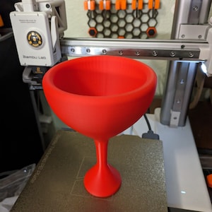 May include: A bright red 3D printed wine glass with a stem and a wide bowl. The glass is sitting on a grey textured surface.