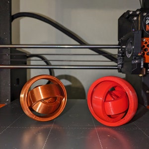 May include: Two 3D printed objects, one red and one copper, are on a black surface. The objects are spherical and have a complex, interwoven design.