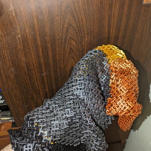 Puede incluir: Un primer plano de un guante de malla hecho de anillos de metal negro, gris, naranja y dorado. El guante está hecho de anillos de metal entrelazados que se tejen juntos para crear un material flexible y duradero.