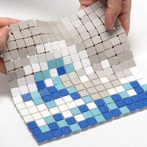 Pode incluir: Uma folha de mosaico flexível segurada por mãos, com um padrão de ondas. O design incorpora azulejos quadrados em tons de azul, branco e cinza. Os azulejos são dispostos para criar um efeito texturizado e tridimensional, adequado para decoração de casa.
