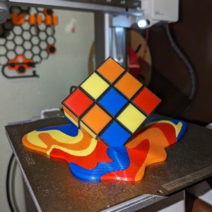 Op de afbeelding: Een 3D-geprinte Rubik's Cube met rode, gele, blauwe, oranje en zwarte vierkanten, die op een 3D-geprinte basis staat met een draaiende patroon van rood, geel, blauw en oranje.