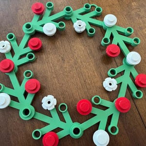 May include: A green plastic wreath with red and white circular accents. The wreath is made of interconnected, stylised branches with circular attachments. The red and white accents are evenly spaced around the wreath, creating a festive appearance.