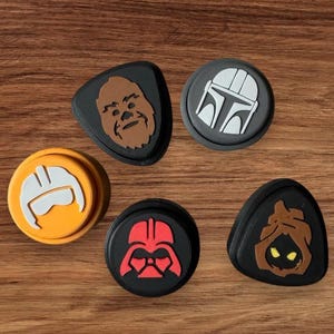 May include: Five different Star Wars character themed controller caps. The caps are made of a soft, flexible material and feature a variety of colours, including black, grey, brown, orange, and red. The characters featured are Chewbacca, Boba Fett, a Stormtrooper, Darth Vader, and a hooded figure.