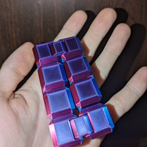 Puede incluir: Un juguete para fidget impreso en 3D con un acabado degradado azul y morado. El juguete está formado por varios cuadrados entrelazados.