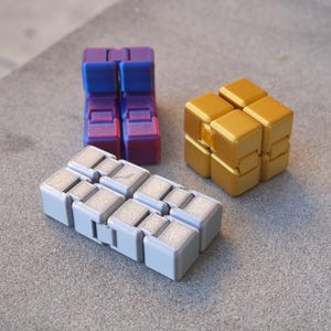 Puede incluir: Tres cubos fidget de diferentes colores. Uno es un degradado de morado y azul, otro es dorado y el tercero es plateado y gris. Los cubos están hechos de pequeños bloques conectados que se pueden manipular.