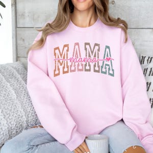 May include: A light pink sweatshirt with the word "MAMA" in a multi-colored, block letter design. Below the word "MAMA" is the word "mama" in a cursive font, with a small pink heart. The sweatshirt is a casual style.
