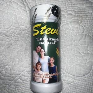 以下が含まれることがあります： ステビア天然甘味料の円筒形容器。ラベルには黄色のブランド名、「Endulzando natural」というフレーズ、家族の肖像画が描かれています。容器は緑と白です。