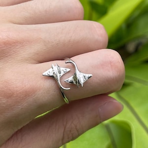 Puede incluir: Un anillo plateado con un diseño de doble manta raya. El anillo se envuelve alrededor del dedo, con las figuras de manta raya en la parte superior. El anillo se encuentra sobre un fondo de follaje verde.