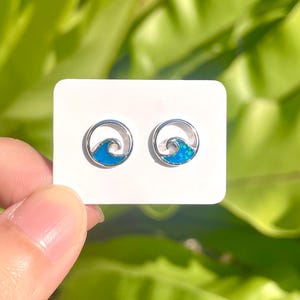 Puede incluir: Pendientes de botón plateados con diseño de ola y centro de esmalte azul. Los pendientes se muestran sobre una tarjeta blanca, con un fondo de exuberante follaje verde.