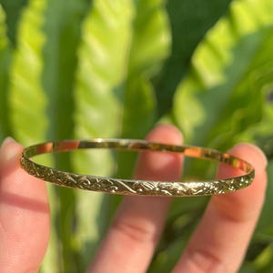 May include: A gold-coloured bangle bracelet with an intricate floral pattern. The thin, circular bracelet is held up against a backdrop of green foliage. The bracelet's design is visible, showcasing its delicate details.