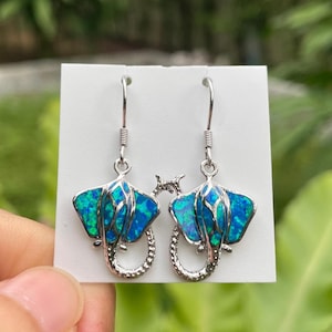 Op de afbeelding: Zilverkleurige oorbellen met een manta rog ontwerp. De oorbellen hebben een blauw en groen opaal inlegsel en een gebogen staart. De oorbellen worden gepresenteerd op een witte kaart.