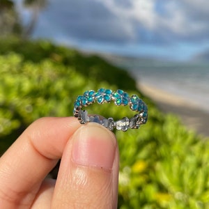 May include: A silver ring with a floral design. The ring features a band of small, turquoise-colored flowers. The ring is held up against a backdrop of green foliage, a beach, and the ocean.