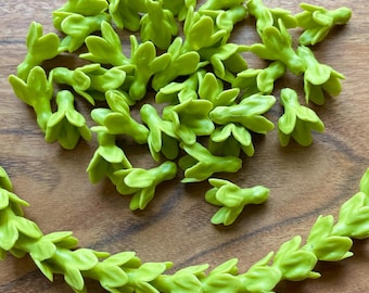 May include: A collection of small, bright green floral embellishments scattered on a wooden surface. Some are loose, while others are arranged in a curved garland shape. The pieces have a delicate, three-dimensional design, suggesting they are for crafting or decorative purposes.