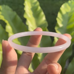 Puede incluir: Un brazalete de jade rosa pálido. El brazalete liso y circular se sostiene sobre un fondo de follaje verde. El color claro del brazalete contrasta con las hojas verdes del fondo.