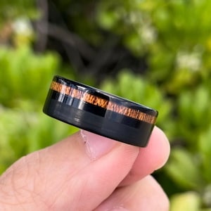 Puede incluir: Un anillo de carburo de tungsteno negro con una incrustación de madera marrón. El anillo tiene un acabado pulido y un ajuste cómodo.