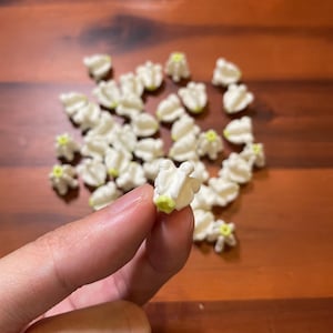 May include: A collection of small, white, flower-shaped candies with a yellow center, scattered on a wooden surface. One candy is held between fingers. The candies have a textured, ruffled appearance.