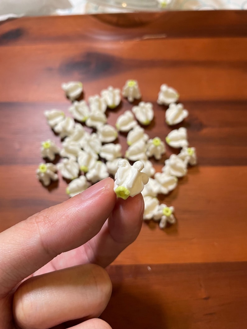 May include: A collection of small, white, flower-shaped candies with a yellow center, scattered on a wooden surface. One candy is held between fingers. The candies have a textured, ruffled appearance.