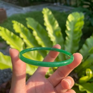 May include: A bright green jade bangle. The circular bracelet is held against a background of green leaves. The jade has a translucent appearance, and the colour is a rich, verdant green.