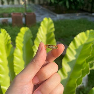 May include: A gold-colored ring with a detailed floral pattern. The ring is held up against a backdrop of green foliage and a blurred outdoor setting. The ring's intricate design is visible.