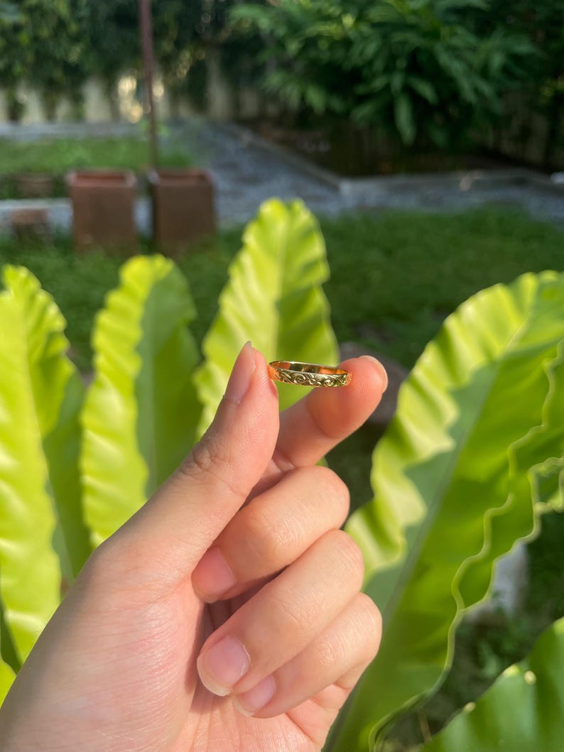 May include: A gold-colored ring with a detailed floral pattern. The ring is held up against a backdrop of green foliage and a blurred outdoor setting. The ring's intricate design is visible.