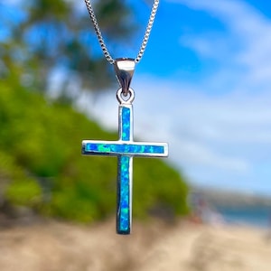 Könnte beinhalten: Halskette mit einem silberfarbenen Kreuzanhänger mit blau-grüner, schillernder Einlage. Das Kreuz hängt an einer filigranen Kette. Der Hintergrund ist eine verschwommene Ansicht von Strand und Himmel.