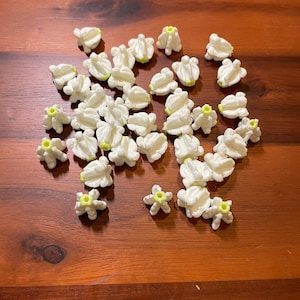 May include: A collection of small, white, flower-shaped embellishments with yellow centers scattered on a wooden surface. The flowers have multiple petals and a textured appearance, suggesting they are made of a fabric or paper material.
