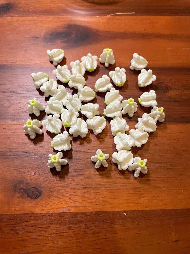 May include: A collection of small, white, flower-shaped embellishments with yellow centers scattered on a wooden surface. The flowers have multiple petals and a textured appearance, suggesting they are made of a fabric or paper material.