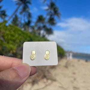 Puede incluir: Pendientes de botón dorados con forma de piña sobre una tarjeta blanca, con un fondo borroso de playa. Los pendientes presentan un diseño texturizado, mostrando un tema tropical. El fondo incluye palmeras, cielo azul y arena.