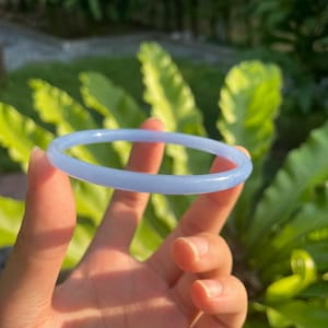 May include: A light blue jade bangle bracelet. The smooth, circular bracelet is held up against a backdrop of green foliage. The bangle's color is a soft, pale blue, and it appears to be made of a translucent material.