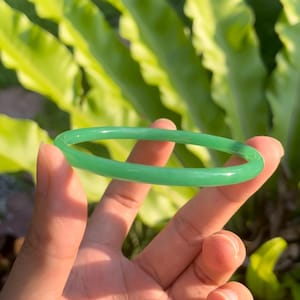 Puede incluir: Un brazalete de jade verde brillante. El brazalete circular se muestra sobre un fondo de follaje verde. El jade es de un color sólido y translúcido. El brazalete tiene aproximadamente 6,35 cm de diámetro.