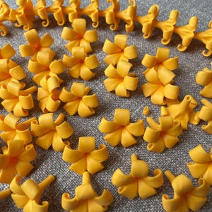 May include: A collection of bright yellow, bow-shaped floral decorations. Some are individual flowers, while others are connected in a chain. The flowers have a detailed, layered design, and are arranged on a gray surface.