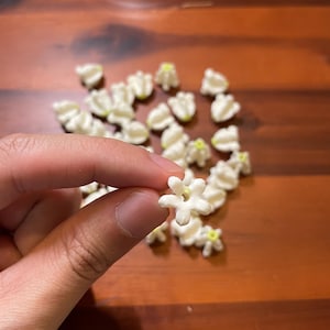 May include: A hand holding a small, white, star-shaped flower with a yellow center. Several similar flowers are scattered on a dark wooden surface. The flowers appear to be made of a soft material.