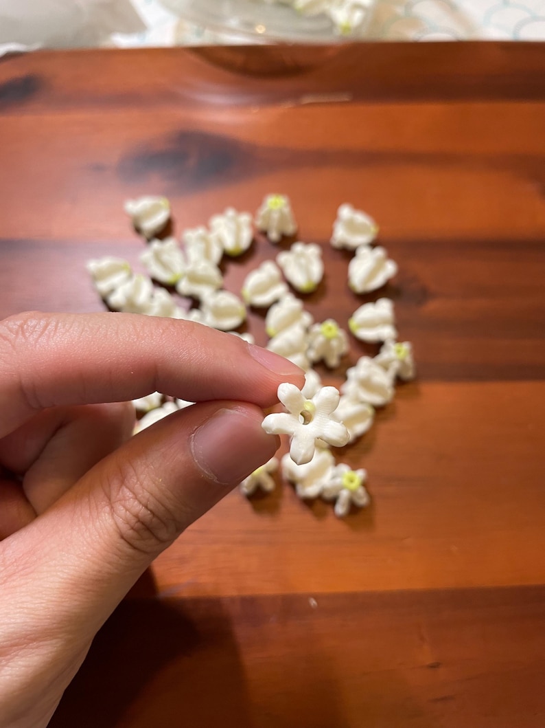 May include: A hand holding a small, white, star-shaped flower with a yellow center. Several similar flowers are scattered on a dark wooden surface. The flowers appear to be made of a soft material.