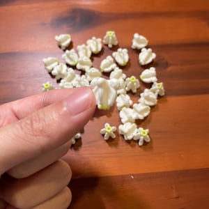 May include: A collection of small, white, flower-shaped decorations with yellow centers. The decorations are scattered on a wooden surface. One decoration is held between fingers.