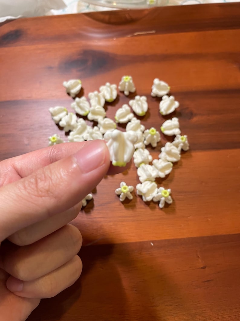 May include: A collection of small, white, flower-shaped decorations with yellow centers. The decorations are scattered on a wooden surface. One decoration is held between fingers.