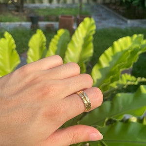 May include: A gold ring with an engraved floral pattern. The ring is worn on a finger, with a blurred background of green foliage. The ring's design features a repeating pattern of leaves and flowers.
